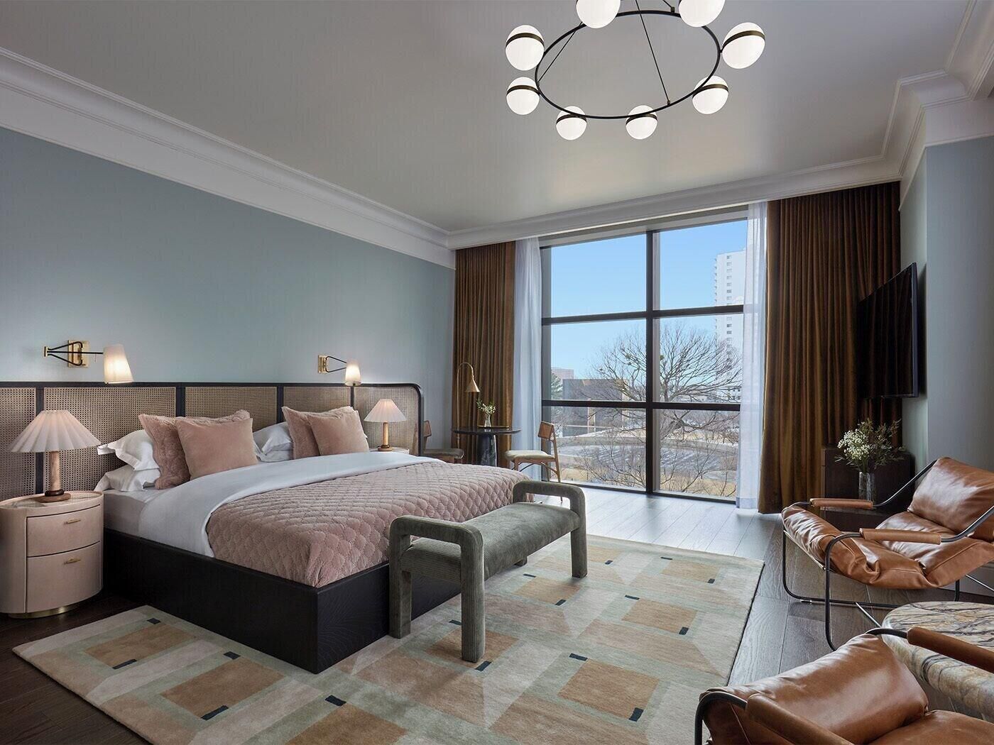 A modern bedroom with a large bed, bedside tables, a bench, a sofa, and a large window offering a city view.
