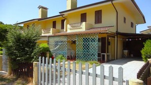 Una casa amarilla de dos pisos con un seto blanco, un porche cubierto y un pequeño jardín.