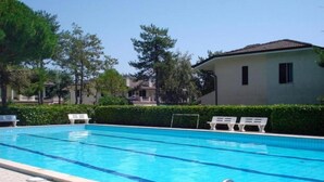 Una piscina con agua azul cristalina, rodeada de sillas reclinables blancas y un seto.