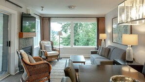 Ein modernes Hotelzimmer mit einer Couch, Sesseln, einem Esstisch, einem Fernseher und großen Fenstern mit Blick auf Bäume.