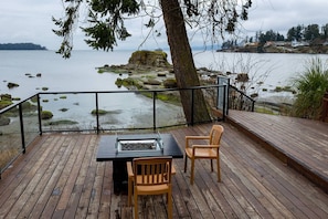 Una terraza de madera con una mesa de vidrio y dos sillas, con vista a una costa rocosa y aguas tranquilas.