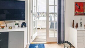 Ein Zimmer mit Fernseher, einem Schreibtisch mit Dekoration, einem großen Fenster mit Blick auf einen Hafen und einer Tür, die zu einem Außenbalkon führt.