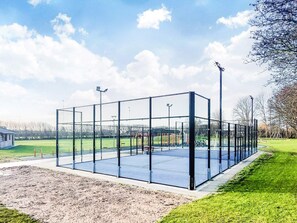 Een tennisbaan met glazen wanden, omgeven door gazon en bomen.