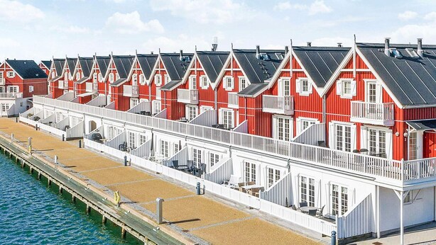 Eine Reihe von roten und weißen Häusern am Meer mit Balkonen, ein hölzerner Steg und ein klarer Himmel.