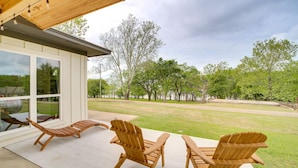 Une terrasse couverte avec trois fauteuils Adirondack en bois et une table, donnant sur une zone herbeuse bordée d’arbres.