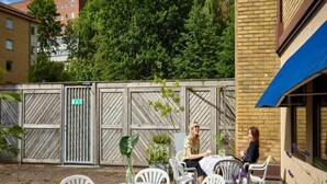 An outdoor seating area with white plastic chairs and tables, a wooden fence, and a building with a blue awning.