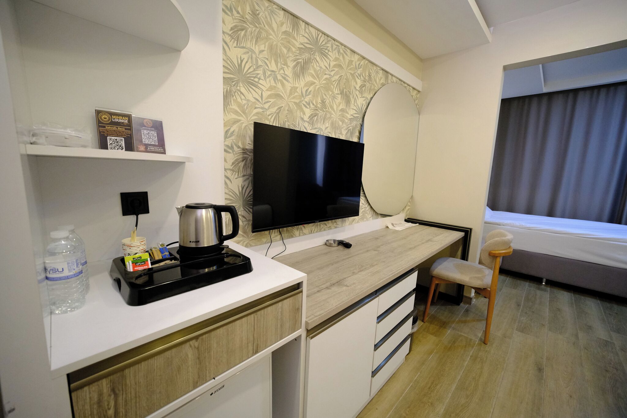 A hotel room with a flat-screen TV, a coffee maker, a water bottle, a small table, a chair, and a window with curtains.