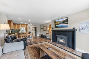 A modern living room with a fireplace, a TV mounted above it, a dining area with wooden furniture, and a kitchen in the background. - Ski in, Ski Out at The Seasons @ Avon (Avon)
