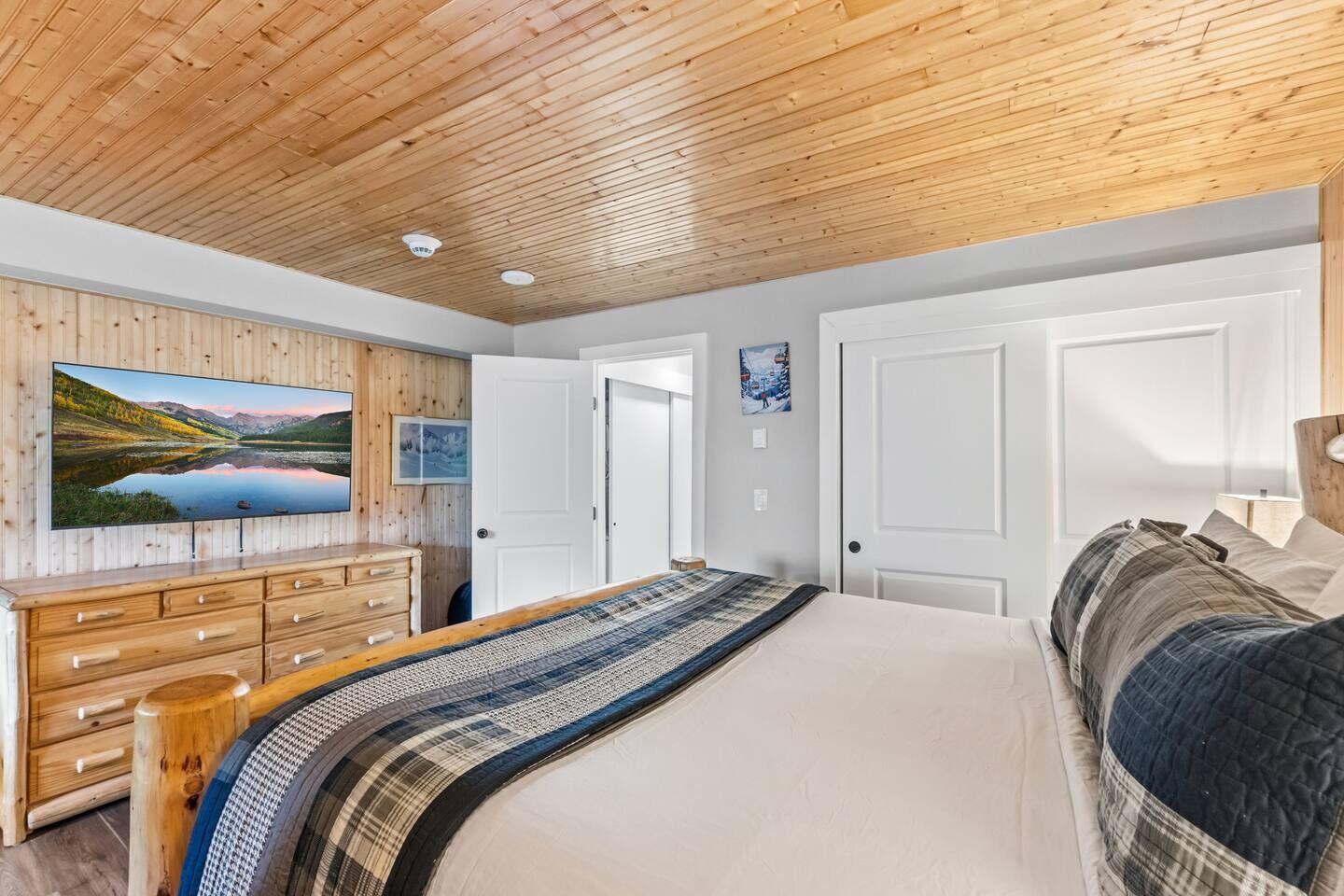 Un dormitorio con techo de madera, una cama grande con colcha estampada, un televisor en la pared y un aparador.