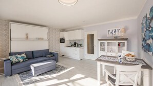A modern living room with a sofa, a dining table with chairs, a kitchen area, and a ceiling light fixture. - Düneneck 21 - lovingly designed one-room domicile (Zempin)