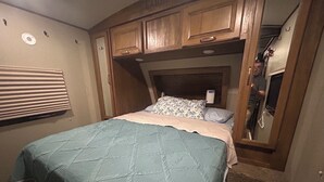 A bedroom with a bed, wooden cabinets, a window with blinds, and a mirror. - Cozy Smoky Mtn RV with WiFi and Heat/AC near Cades Cove (Walland)