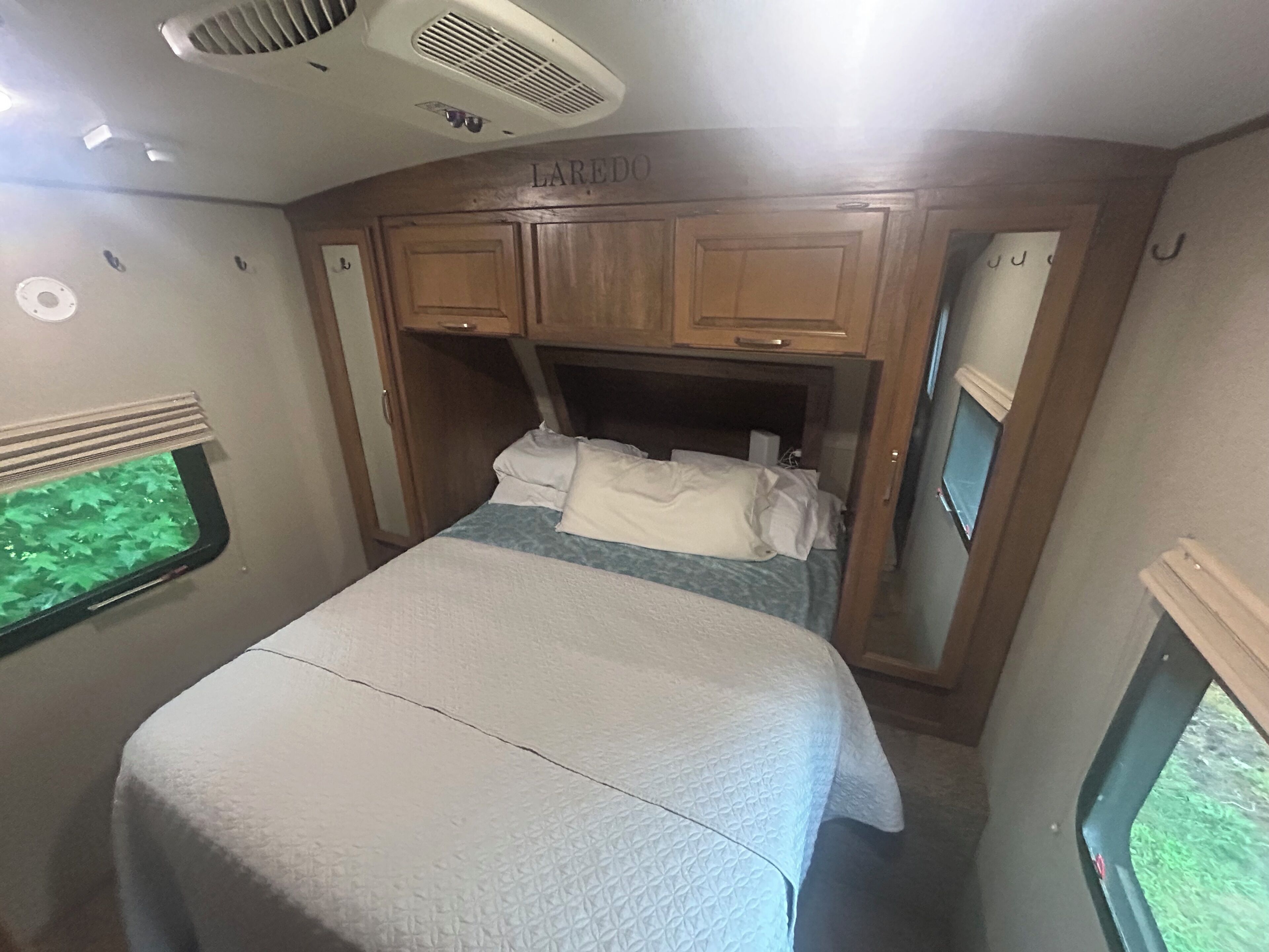 Un dormitorio de autocaravana compacto con cama, cabecero, ventana y respiradero en el techo.