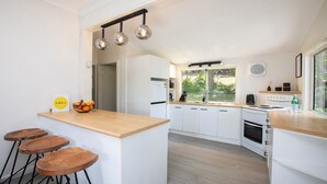 A modern kitchen with white cabinets, a wooden countertop, and a fruit bowl. There are three stools and a hanging light fixture.