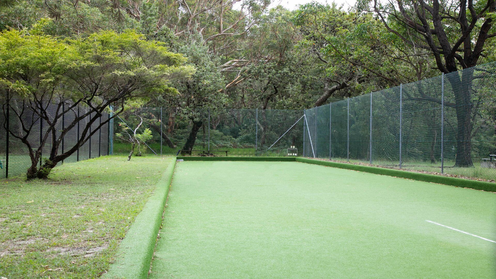 A green artificial turf tennis court surrounded by trees and a chain-link fence.
