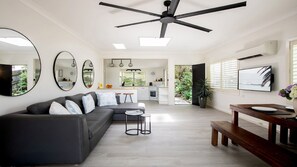 A modern living room with a dark sofa, a coffee table, and a TV. There is a kitchen in the background with a window and a plant. - Trampoline + 150mtrs to beach (PEARL BEACH)