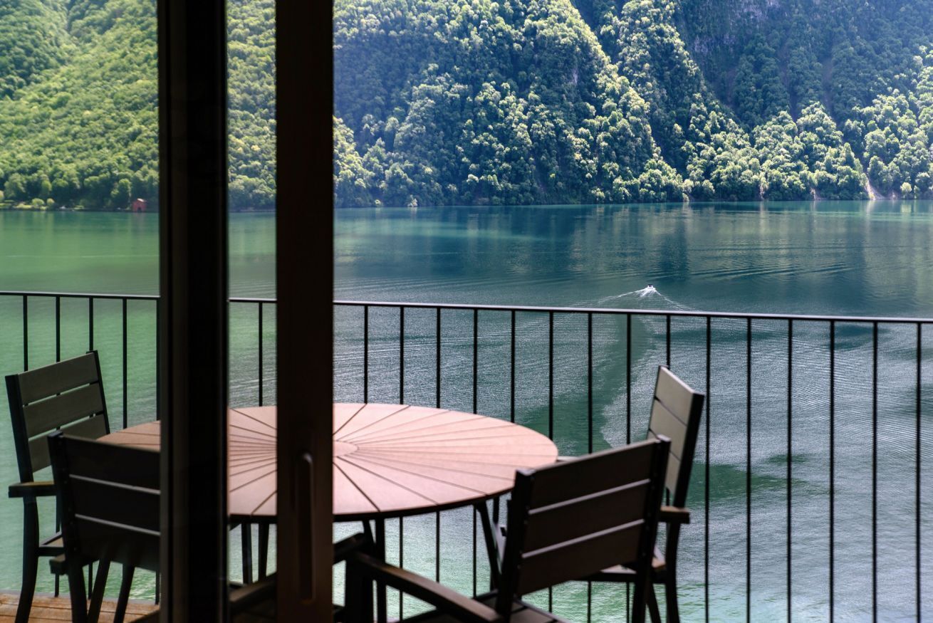 Un balcon avec une table et des chaises donnant sur un lac, avec une colline boisée en arrière-plan.