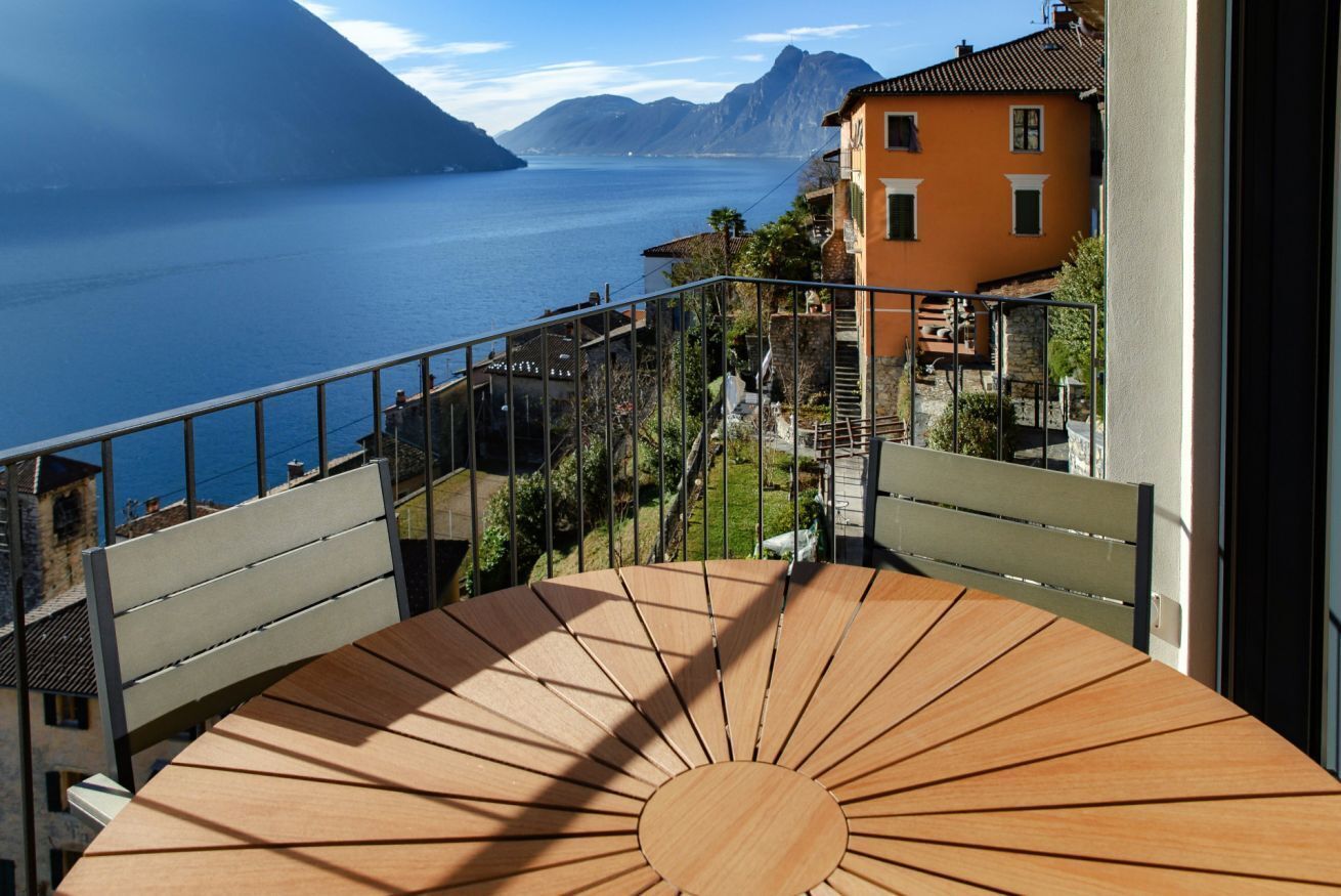Un balcón con mesa y sillas de madera, con vistas a una zona costera con casas y una extensión de agua.