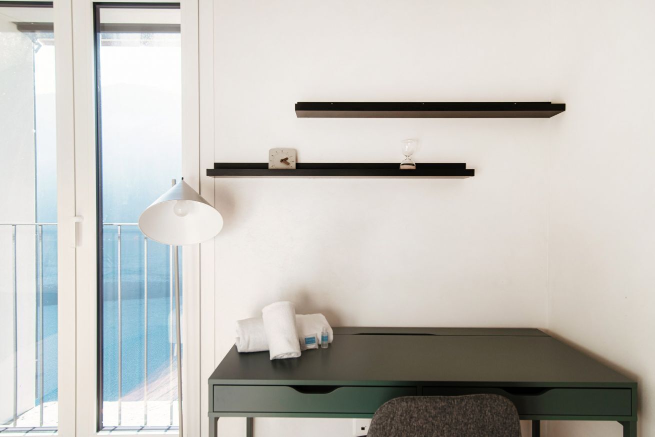 A room with a desk, a chair, a lamp, and two shelves with a clock and a glass.