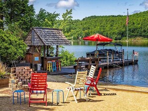 A lakeside dock with a small wooden structure, a red umbrella, and several colorful chairs.