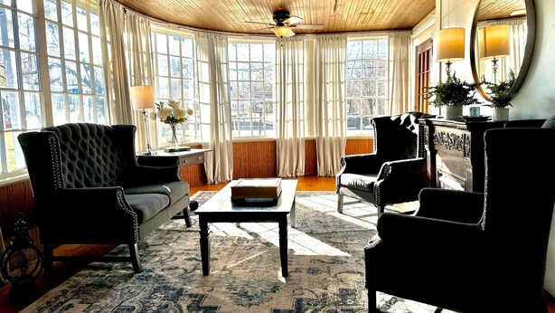A room with a wooden ceiling, large windows with curtains, a black tufted armchair, a sofa, a coffee table, and a mirror on the wall. - Home Near Hiking Biking w Hottub 6 Bedrooms (Bennington)