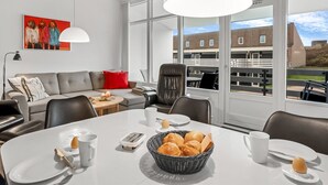 Ein modernes Wohnzimmer mit einem großen Fenster, einem gedeckten Esstisch mit Brot und einer Kaffeetasse, einer roten Couch mit Kissen und einem Gemälde an der Wand.