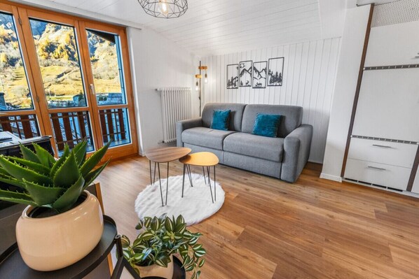 A modern living room with a grey sofa, a small coffee table, and two chairs. There are potted plants on the floor, a white rug, and a window with a view of a mountainous landscape. - Beautiful, centrally located apartment in Leukerbad (Leukerbad)