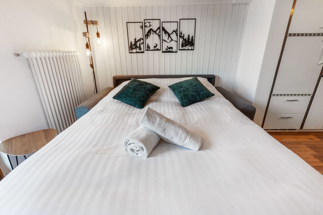 En pænt redt seng med to grønne puder og et sammenrullet hvidt håndklæde, en radiator, samt en væg med fire billeder i rammer.