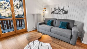 A cozy living room with a grey sofa, two wooden side tables, and a view of a mountainous landscape through a glass door. - Beautiful central apartment in Leukerbad (Leukerbad)
