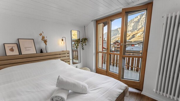 A bedroom with a large bed, wooden headboard, a window with a view of mountains, a radiator, and a bedside table with a lamp. - Beautiful central apartment in Leukerbad (Leukerbad)