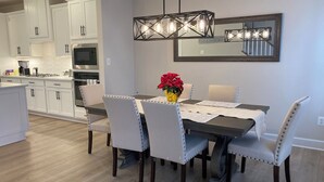 A modern kitchen with white cabinets, a dining area with a dark table and white chairs, and a decorative light fixture above the table. - Spacious, New 4-bdrm townhome in charming Lewes, walk or bike to town & beaches! (Lewes)