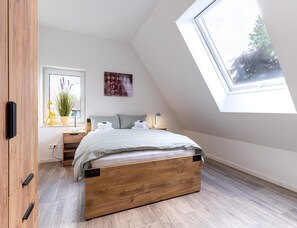 A bedroom with a sloped ceiling, a bed, a bedside table, a lamp, a window with a view, and a door.