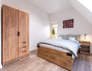 A bedroom with a large wooden wardrobe, a bed with a grey headboard, a nightstand, and a window with a view of the outside. - Ferienhaus in Tating (Tating)