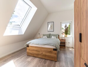 A bedroom with a sloped ceiling, a bed with a wooden frame, a bedside table, a nightstand, a framed picture on the wall, and a window with a view of greenery. - Ferienhaus in Tating (Tating)