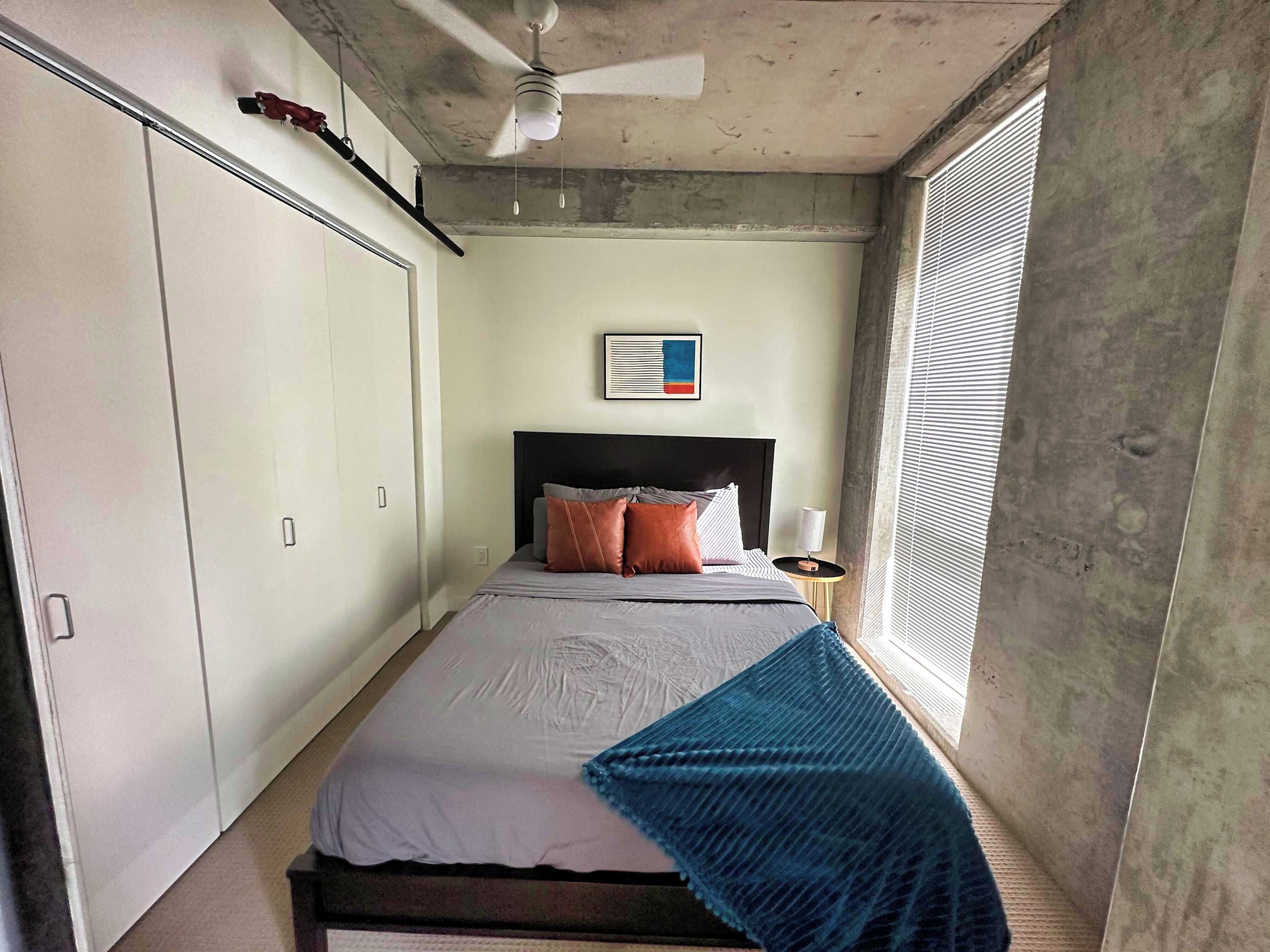 Un dormitorio con una cama, ventilador de techo, ventana con persianas y una obra de arte colgada en la pared.