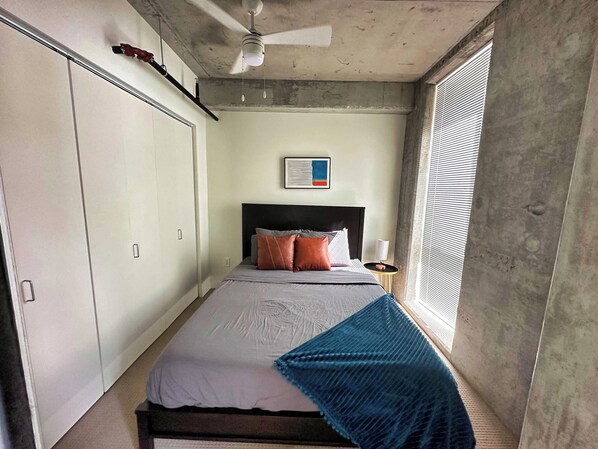 A bedroom with a bed, a ceiling fan, a window with blinds, and a wall-mounted artwork.