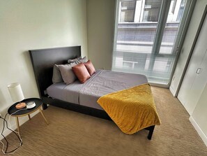 A bedroom with a bed, a bedside table, a lamp, and a window with blinds.
