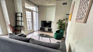Een moderne woonkamer met een grijze bank, een klein bijzettafeltje, een groene fauteuil, een televisie en een potplant.
