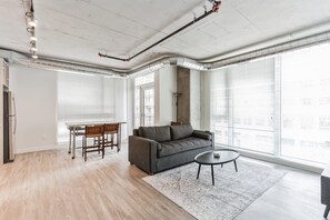 A modern living room with a grey sofa, a round coffee table, and a dining table with chairs. There are large windows with blinds, a ceiling with exposed ductwork, and a light fixture. - 7 Combined units with Stunning City Views (Cleveland)