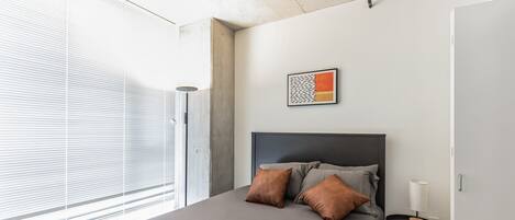 A modern bedroom with a large bed, a bedside table with a lamp, and a window with blinds.