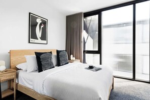 A modern hotel room with a large bed, a nightstand, a framed artwork, and a view through a large window.