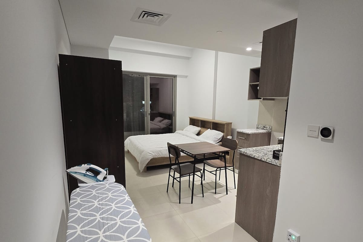 A compact hotel room with a bed, a small dining table with chairs, a kitchenette, and a sliding door leading to a balcony.