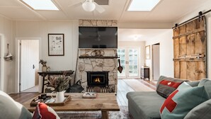 A cozy living room with a stone fireplace, a large flat-screen TV, and a rustic wooden barn door. - Lola Cottage - Your Charming Retreat in the Heart of Adaminaby (Adaminaby)