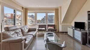 A modern living room with a leather sofa, a glass coffee table, two armchairs, a TV, and a view of the city through large windows.