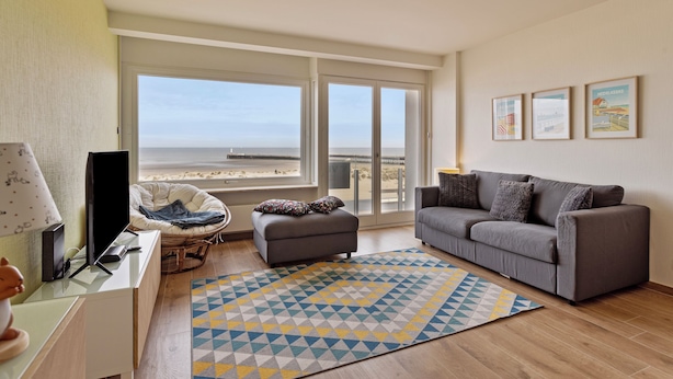 Un salon moderne avec un canapé gris, un petit pouf, un fauteuil en osier et une grande fenêtre donnant sur la plage.