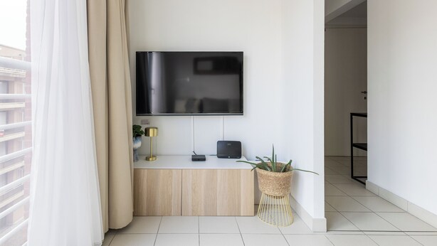 Un soggiorno moderno con una TV a schermo piatto montata sulla parete, un tavolo consolle in legno con una lampada e una pianta, e vista sul balcone esterno.