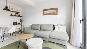 Un soggiorno moderno con un divano grigio, sedie bianche, un tavolino rotondo in legno e un piccolo tavolino bianco. Si nota un termosifone, una mensola con oggetti decorativi e uno specchio appeso al muro.