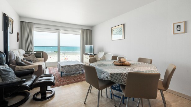 A modern living room with a view of the beach, a dining area with a table and chairs, and a television on a stand.