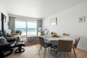 Een moderne woonkamer met uitzicht op het strand, een eethoek met tafel en stoelen, en een televisie op een meubel.