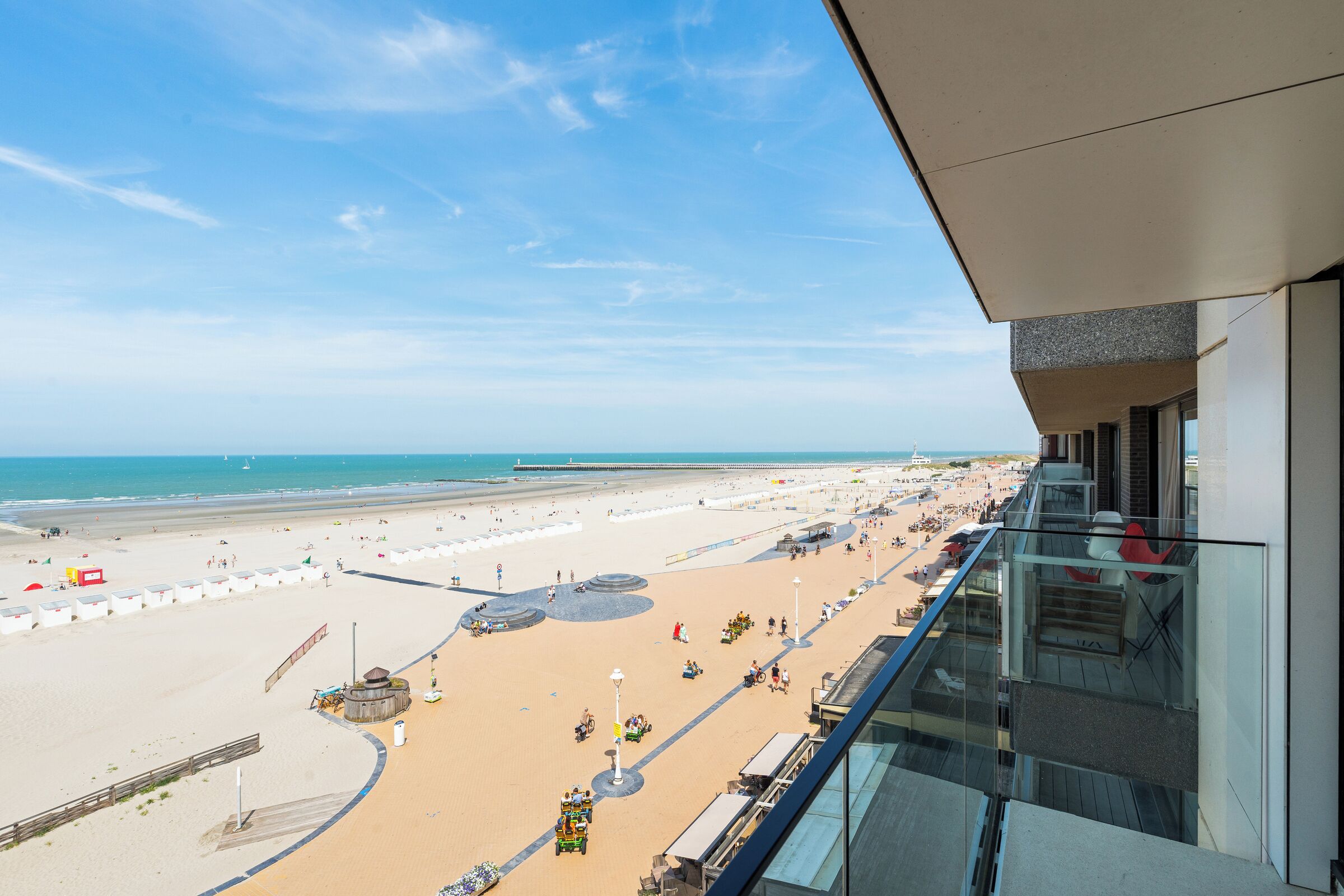 Blick auf einen Strand vom Balkon aus, mit blauem Himmel, Sandstrand und einer Uferpromenade mit Menschen und Badegästen.