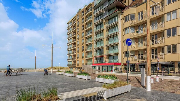 A multi-story hotel with balconies, a red awning, and a pedestrian area with planters and benches. - 2 bedroom accommodation in Nieuwpoort (Nieuwpoort)